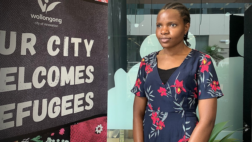 A young African woman stands looking off camera inside a library next to a sign that says 'Our city welcomes refugees'.