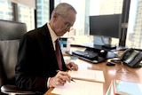 ACCC chairman Rod Sims sits at his desk with a pen marking up documents.