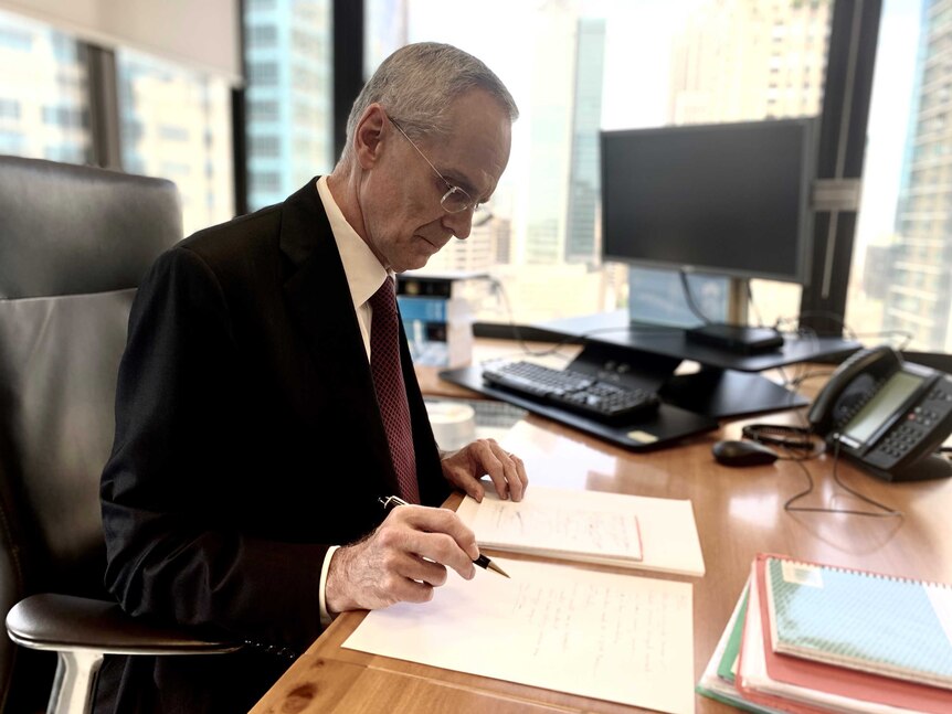 ACCC chairman Rod Sims sits at his desk with a pen marking up documents.