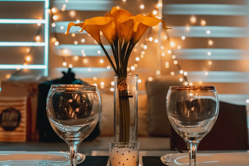 Two wine glasses on a table with a vase of yellow flowers in between
