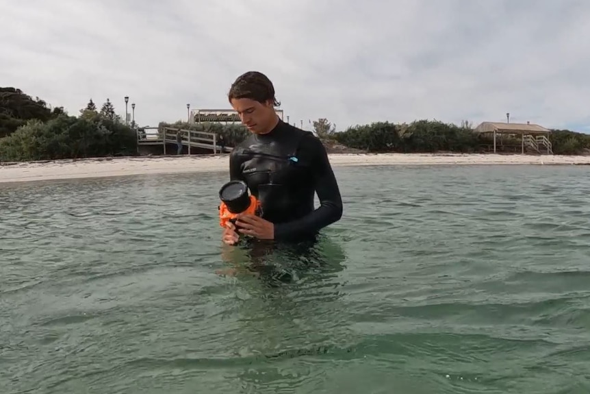 A teenage boy in the ocean with a wetsuit, holding a camera. 
