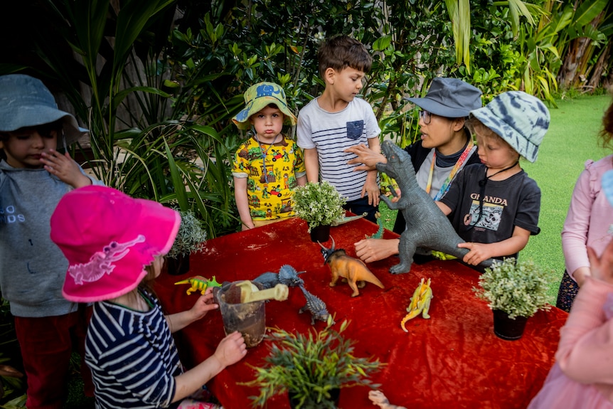 A group of young kids at a childcare centre standing around a table with toy dinosaurs 