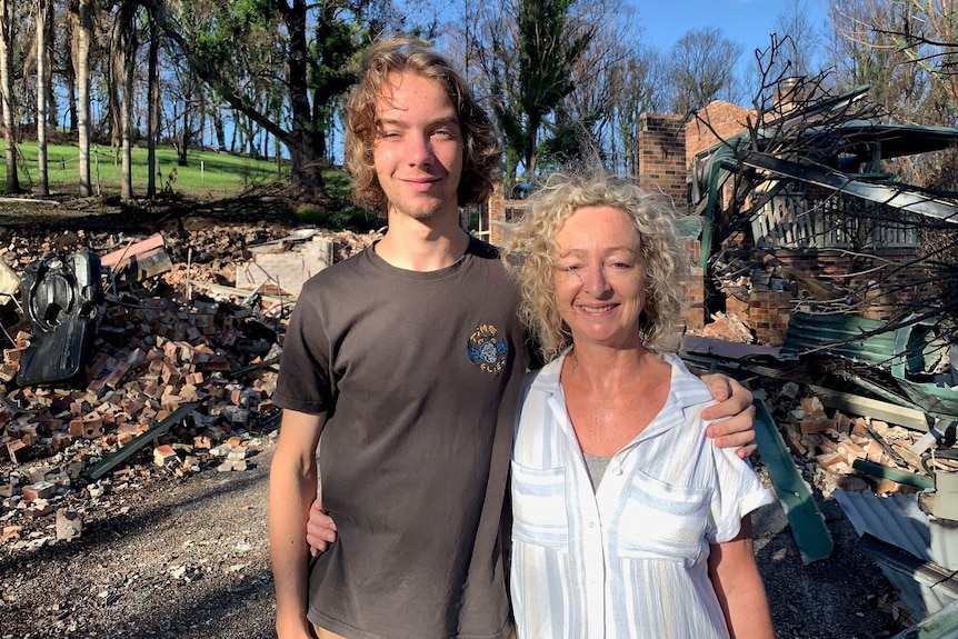 a male teenager with his arm around his mother standing in front of a destroyed home