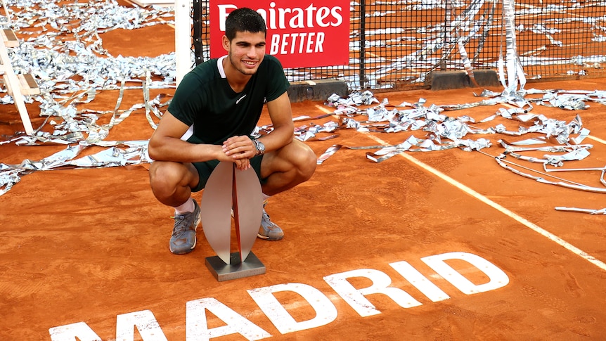 A young tennis star crouches next to the red clay court with the sign 'Madrid' as he holds a tournament trophy.