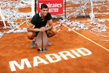 A young tennis star crouches next to the red clay court with the sign 'Madrid' as he holds a tournament trophy.