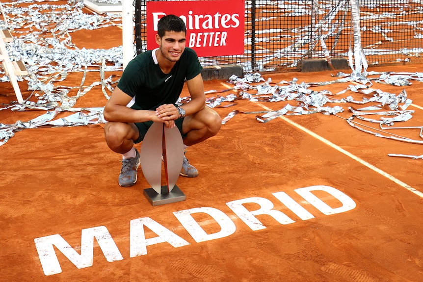 A young tennis star crouches next to the red clay court with the sign 'Madrid' as he holds a tournament trophy.