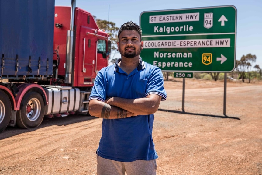 Man with tattooed arms stands in front of road sign, beside a truck