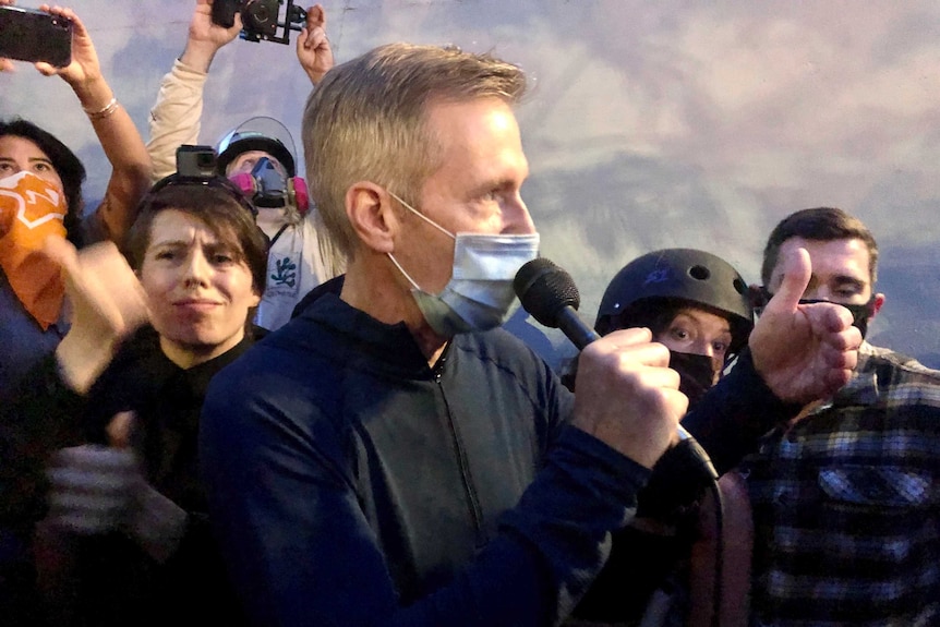 A man in a mask holds a microphone to his face as he addresses a crowd of people around him
