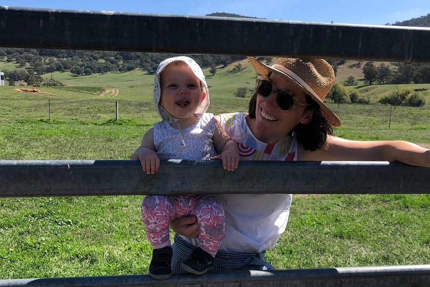 A smiling, dark-haired woman in a hat and sunglasses supports her beaming toddler daughter as she hangs on to a farm fence.