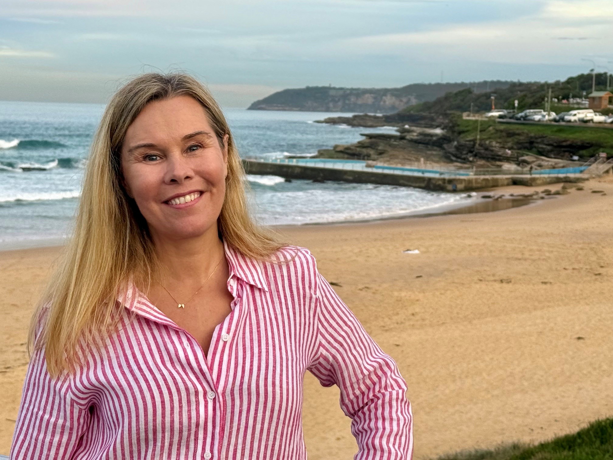 Woman with blonde hair standing in front of a beach.