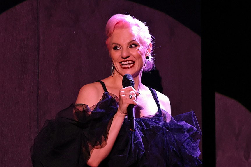 A 30-something trans woman with pink hair, wearing a black dress, talks into a microphone while illuminated by a spotlight