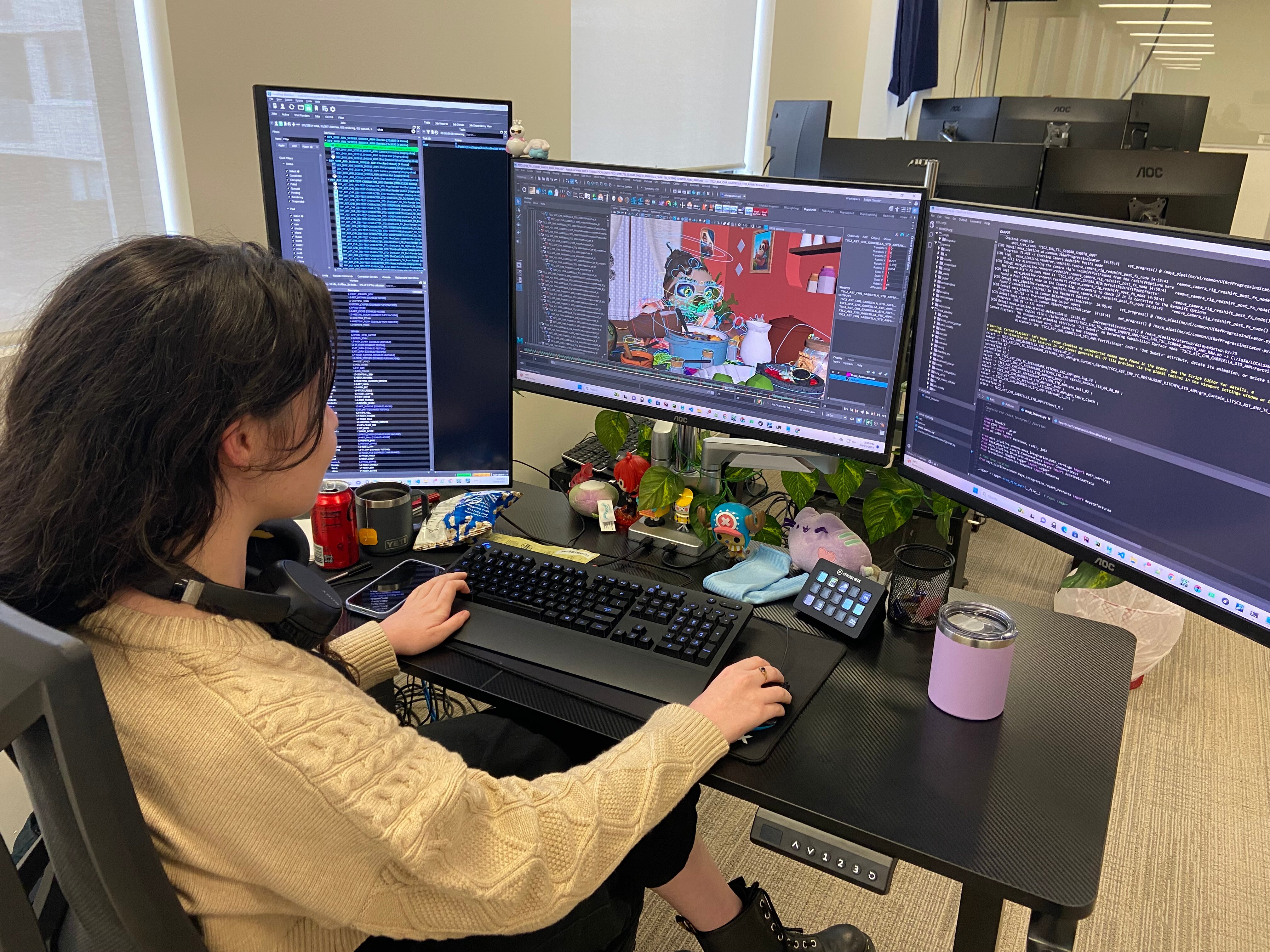 A woman working on a character at a desk, in front of a screen showing an animation and two other screens of code.