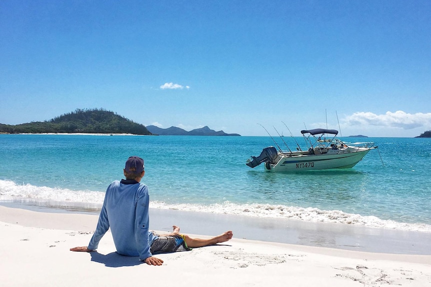 Matt Dillon sits on a white sand beach looking at a boat on bright blue water. 