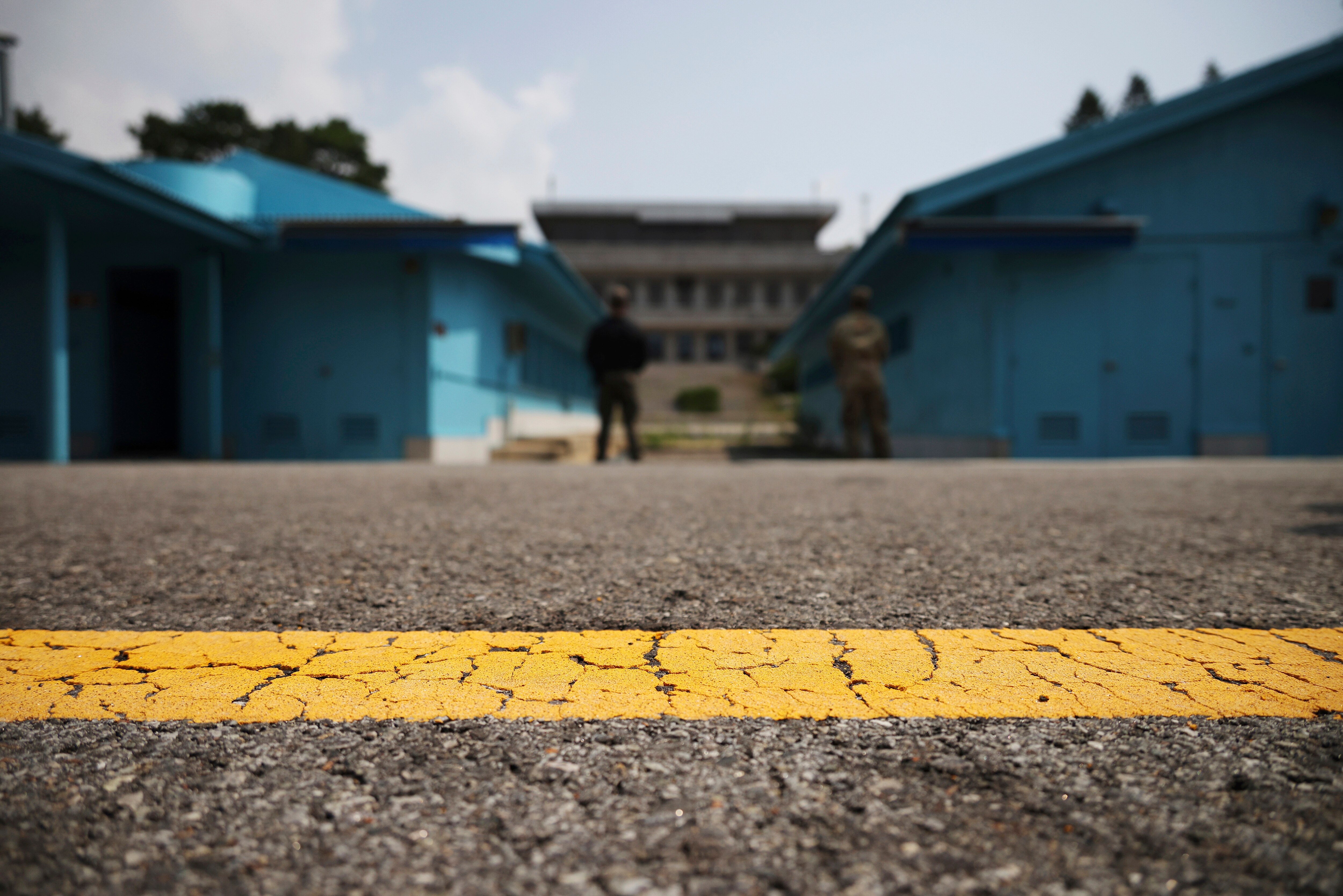 A cracked yellow line runs across an asphalt road, with low blue buildings and soldiers in the background.