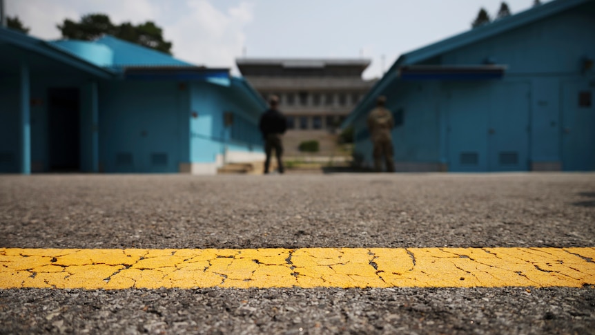 A cracked yellow line runs across an asphalt road, with low blue buildings and soldiers in the background.