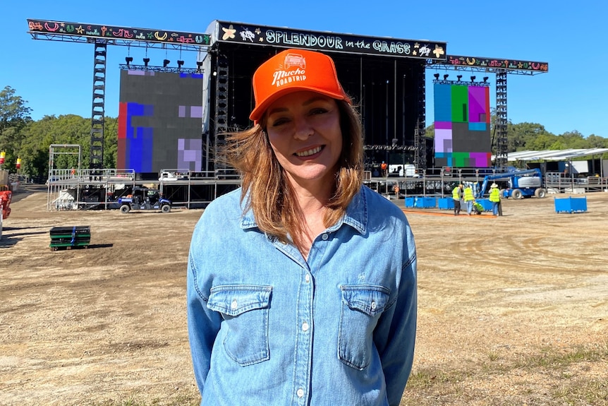 Una mujer con una gorra naranja sonríe y mira directamente a la cámara, se para frente a un escenario musical.