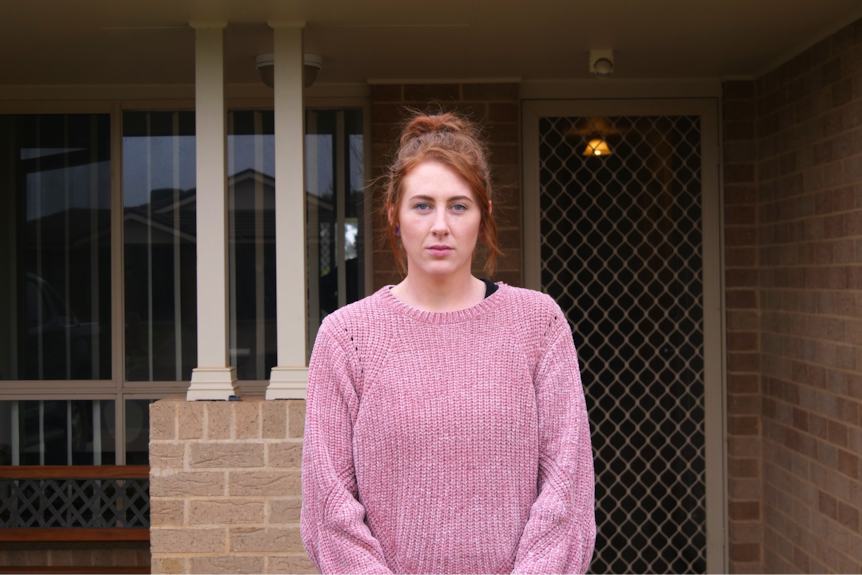 A serious woman with red hair and pink jumper stands in front of suburban home.