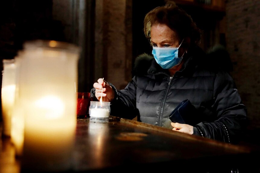 A woman wearing a face mask lights a candle in a dark church.