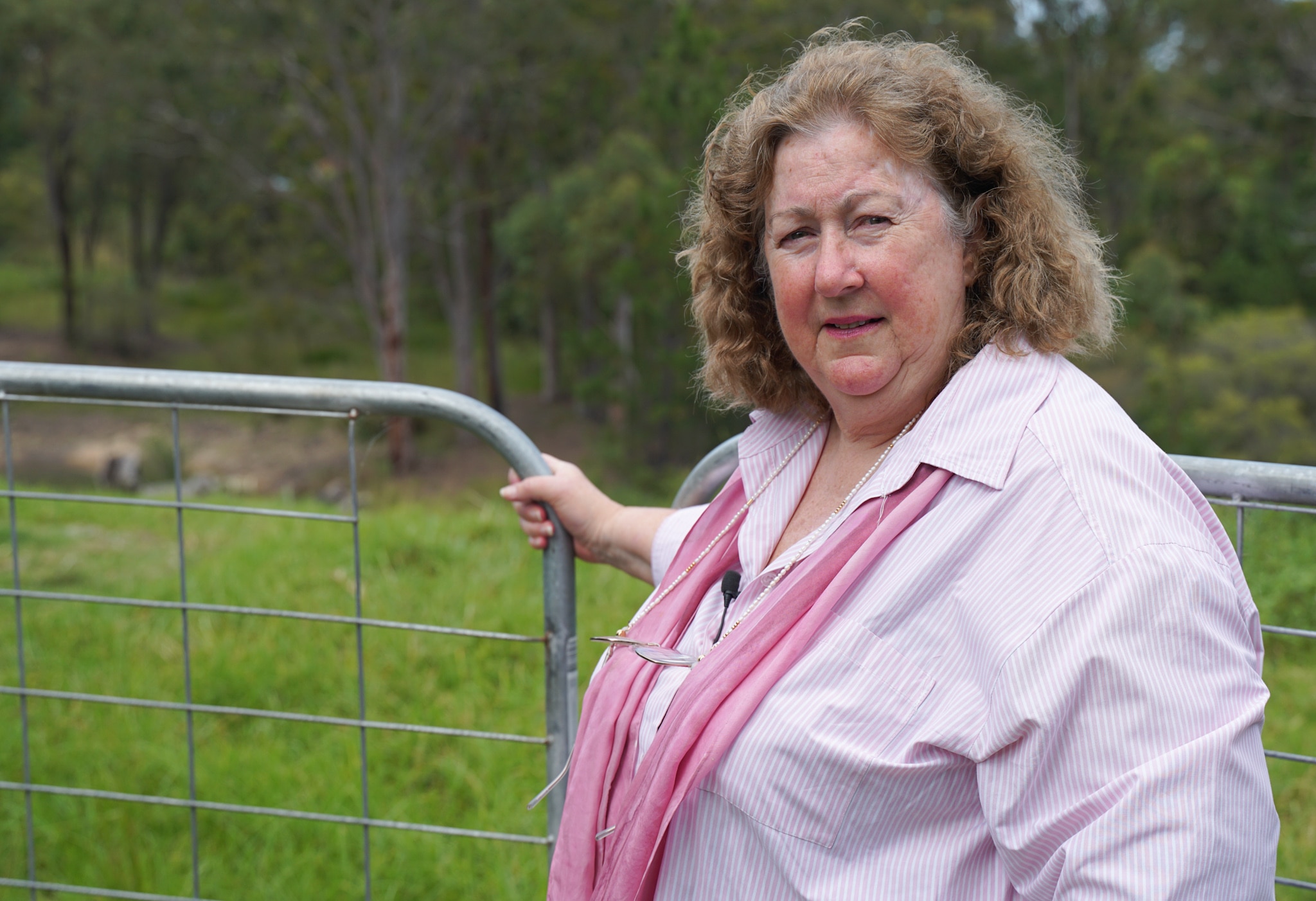 Woman wearing a pink shirt and scarf stands opening farm gate, with green rural setting in the background