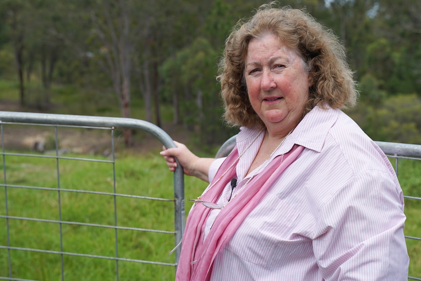 Woman wearing a pink shirt and scarf stands opening farm gate, with green rural setting in the background