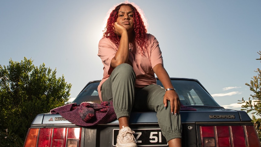Indigenous woman sits atop a blue mitsubishi, she wears pink tshirt and olive coloured pants, sneakers