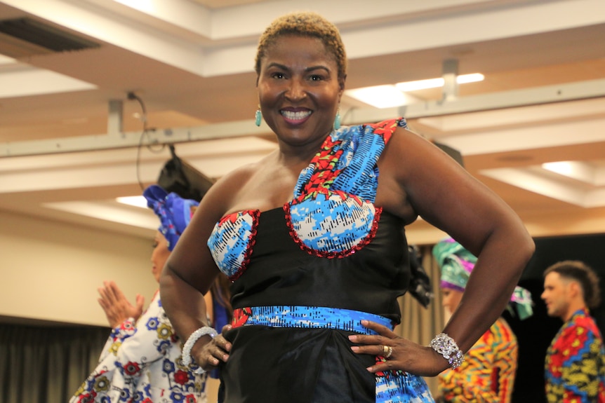A woman poses for a photo on the catwalk at a fashion show wearing a black dress marked with blue and orange patterns.