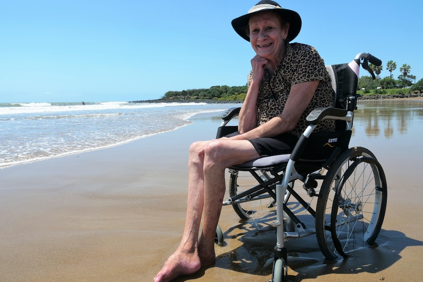 An elderly woman in a beach wheelchair and black sunhat on a beach. 