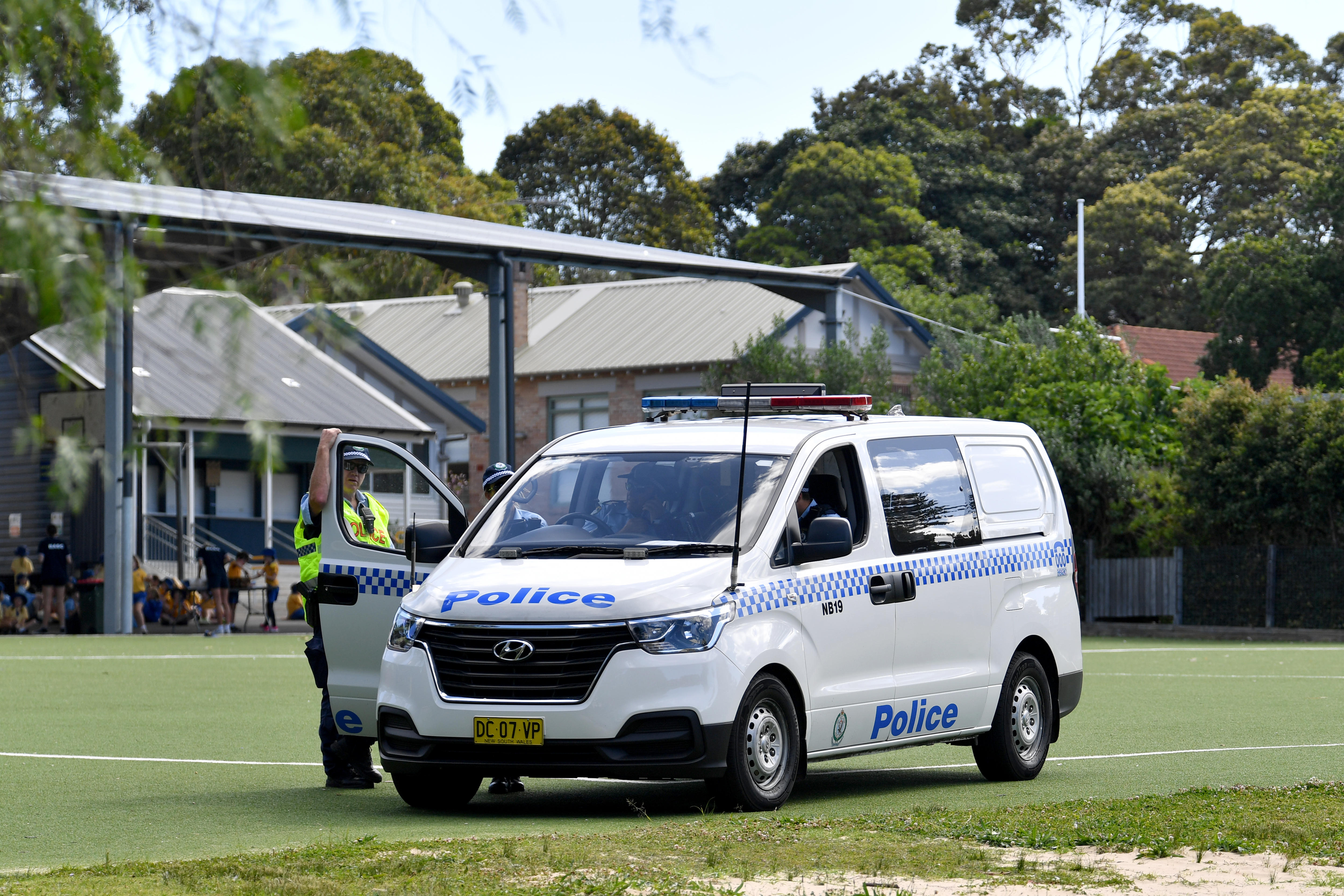 a police officer leans on the door of a police van parked in the middle of a school oval