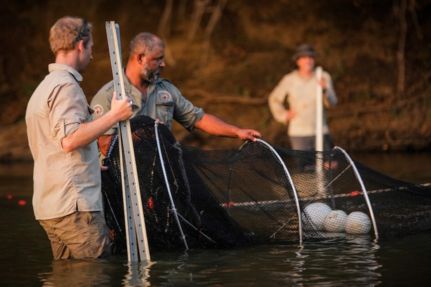 Two men in khaki clothing standing in a river with a large net. The net has plastic rings to hold its shape.