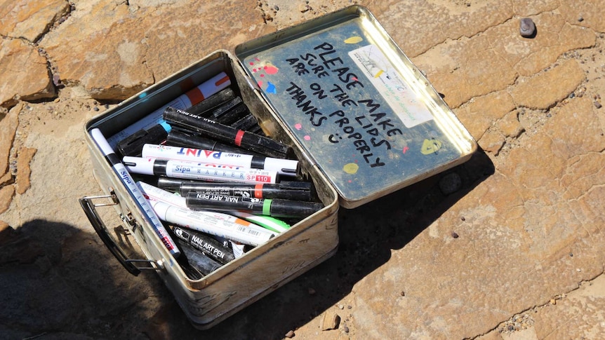 A box full of permanent markers sits on a slab of rock. "Please make sure the lids are on properly thanks :)" is on lid.