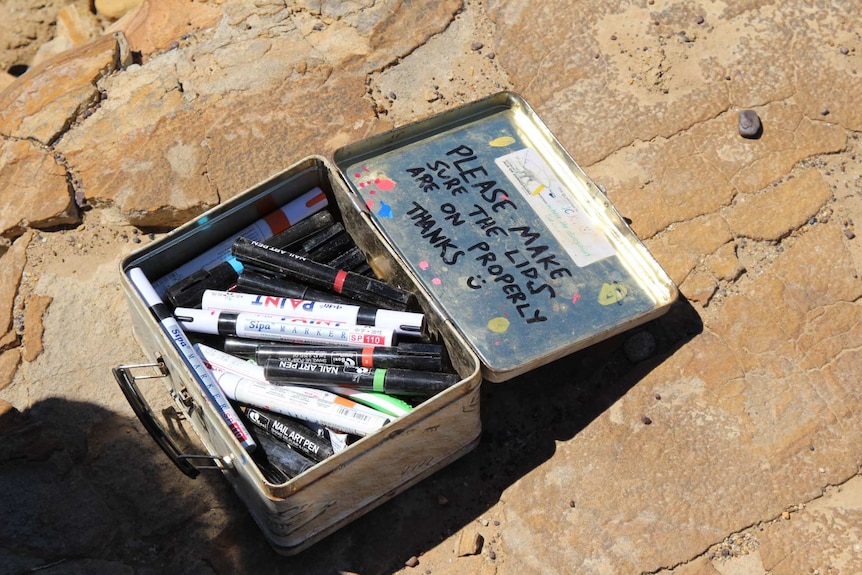 A box full of permanent markers sits on a slab of rock. "Please make sure the lids are on properly thanks :)" is on lid.