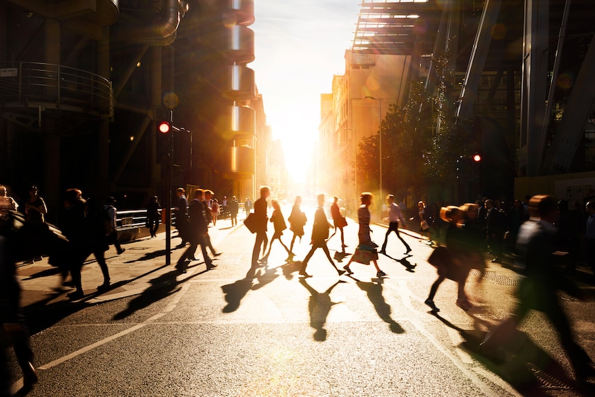 Silhouettes of office workers crossing intersection, sun setting between highrises behind them.