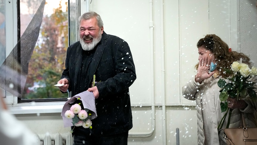 Colleagues spray champagne toward Dmitry Muratov who smiles while holding a bunch of flowers.