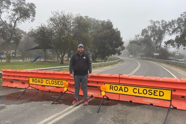 A man in dark clothes and a hat stands in font of a barricade that says "road closed".