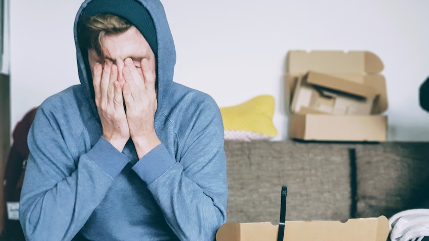 Man in a hoodie sitting on a couch with his hands over his eyes