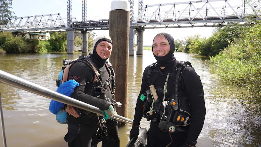 Two divers stand in front of a river in the Hunter region in NSW with the Hinton bridge in the background.