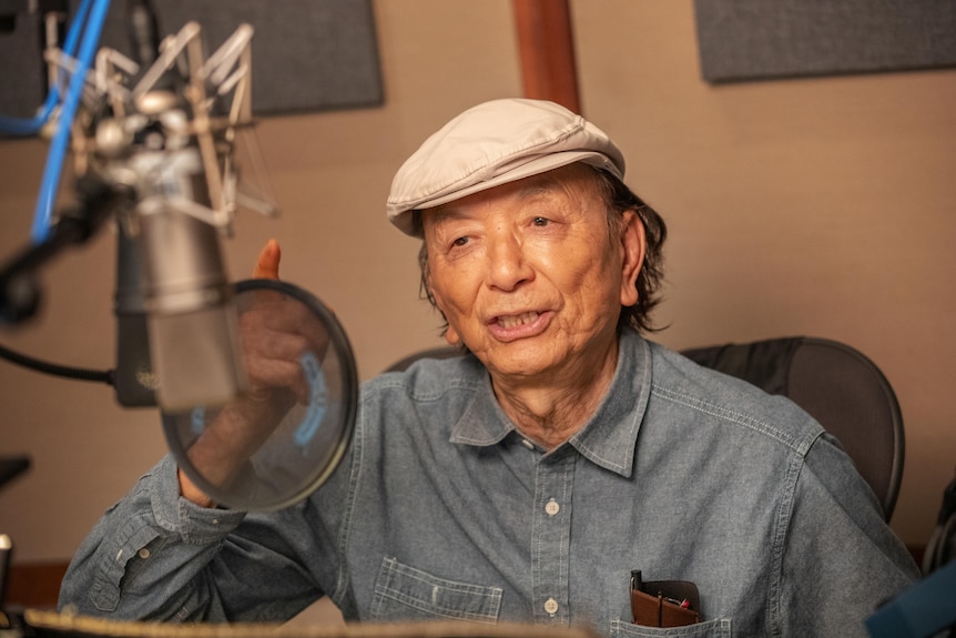 An older man sits talking and gesticulating into a microphone inside a recording studio.