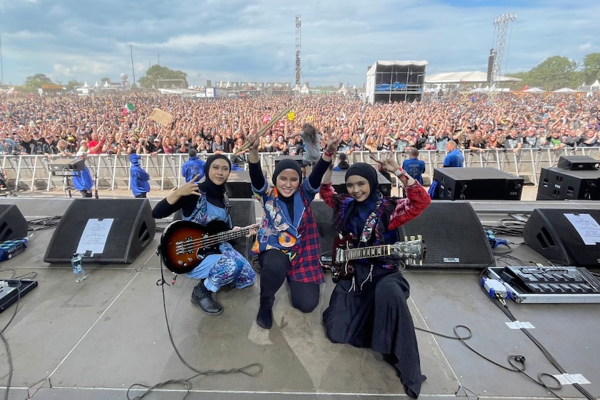 three female hijab wearing rockstars kneel down on a stage with a huge crowd at a rock music festival behind them.