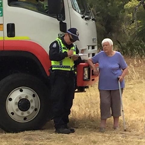 A police officer and an elderly woman with a walking stick stand next to a truck as the officer writes a ticket.