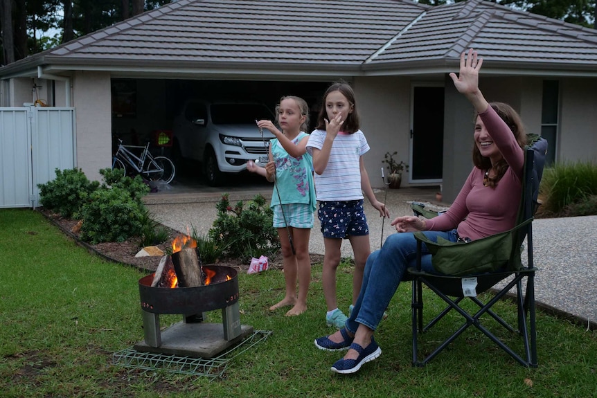 A lady sits on a seat on her front lawn waving, next to a fire pit.