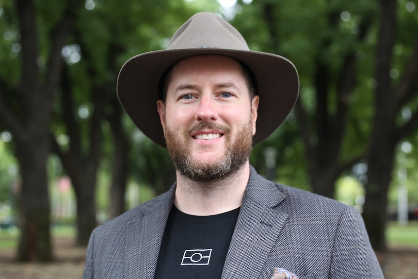 Marcus Stewart, wearing an Akubra and black t-shirt with an Aboriginal flag design, stands in a green park.