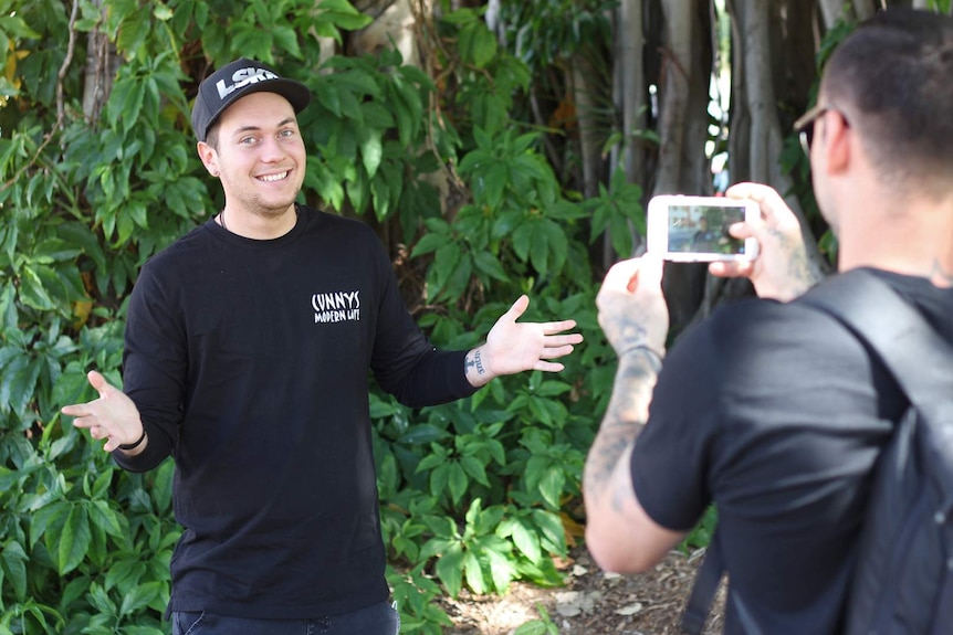 Comedian Josh Wade stands in front of a tree making a funny face while another man takes his photo on a smartphone