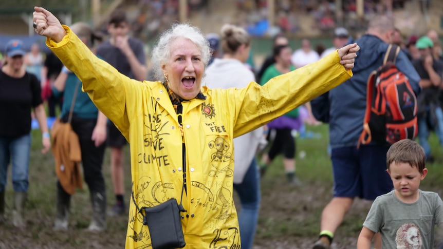 A woman in yellow coveralls dancing on muddy ground with her arms in the air.