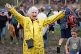 A woman in yellow coveralls dancing on muddy ground with her arms in the air.