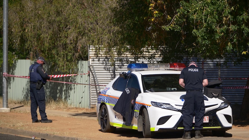 Two police stand by a police car. They are wearing uniforms and both working. There is police tape behind them.