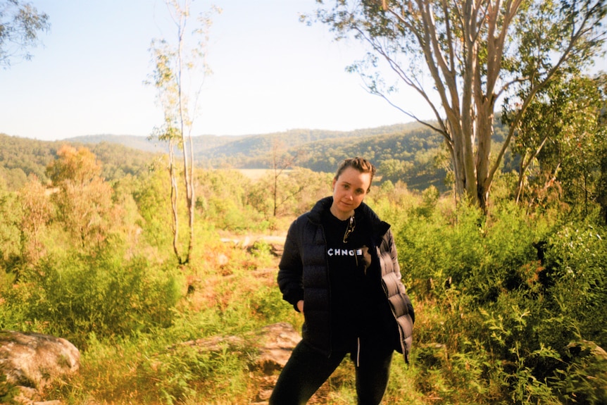 A woman with a serious expression wearing black activewear stands in front of a bushy landscape with mountains in the background