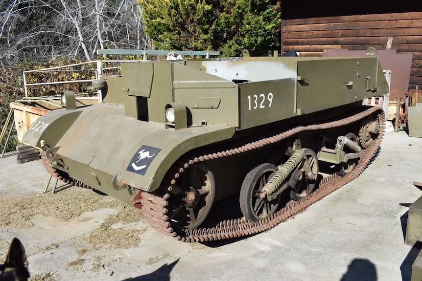 A large, khaki-green military vehicle on treads, sits stationary on a slab of concrete. It is surrounded by machine parts.