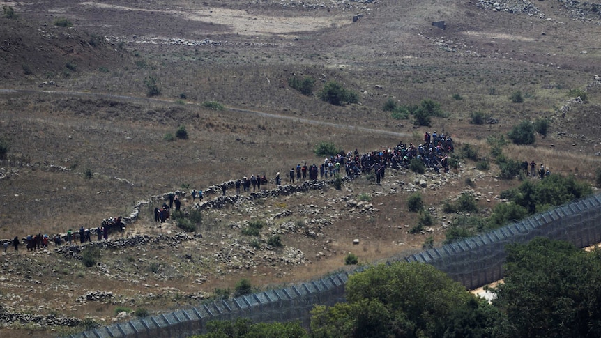 A wide shot shows a group of people in the distance approaching a border fence.