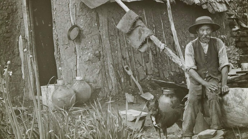 A scan of a black and white image shows a Chinese man seated out front of a mud hut with a dog standing beside him.