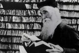 Black and white photo of a man with long beard and glasses, holding a book and surrounded by shelves of many tiny paper slips.